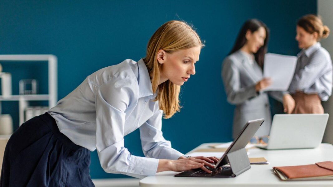 Three things we’ve learnt about the UK economy from the IMF A female office worker wearing a light blue top and dark blue skirt leans over a laptop computer. Two other workers are in the background, slightly out of focus
