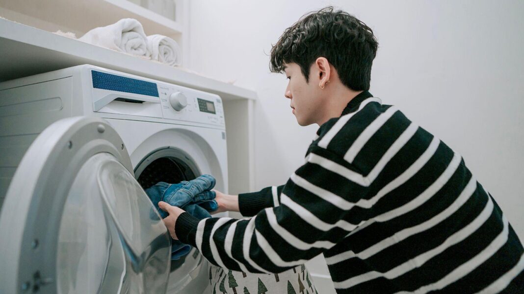 Stock photo shows a person wearing a black and white striped top loads clothes into a washing machine in a laundry room at home.