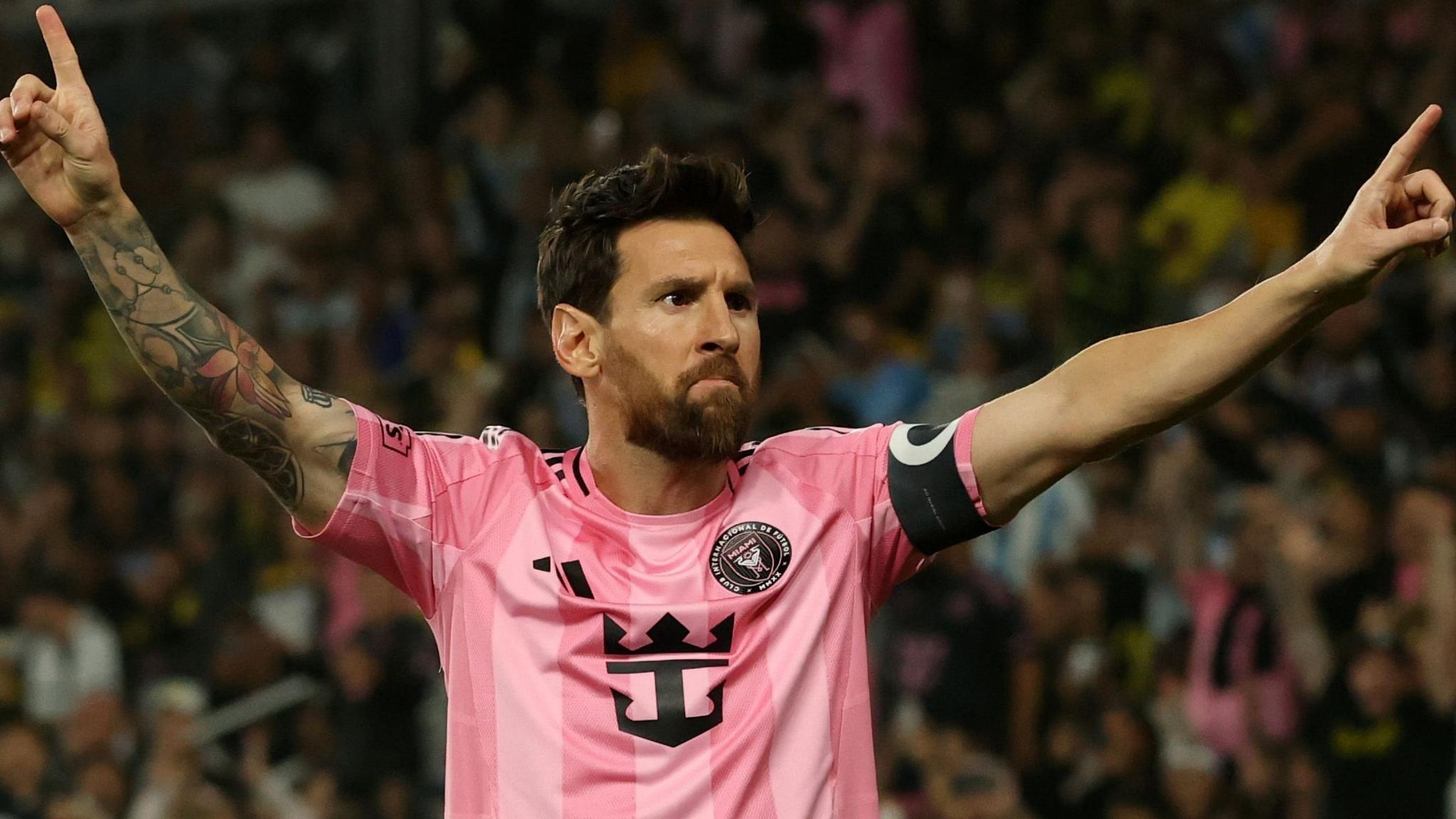Lionel Messi celebrates while playing for Inter Miami in the MLS, wearing their pink kit and stretching out his arms upwards with index fingers pointing