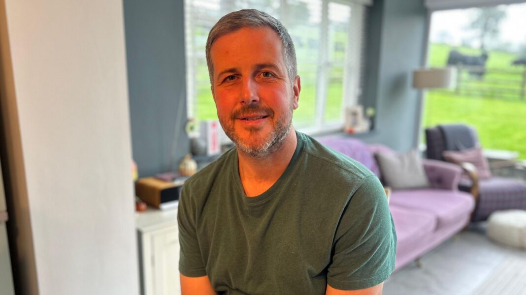 Wales plans to ban steel pylons as government launches consultation A man wearing a green shirt and looking at the camera. Behind him is a purple sofa inside a living room with painted blue/grey walls. A back garden can be seen through the French windows.