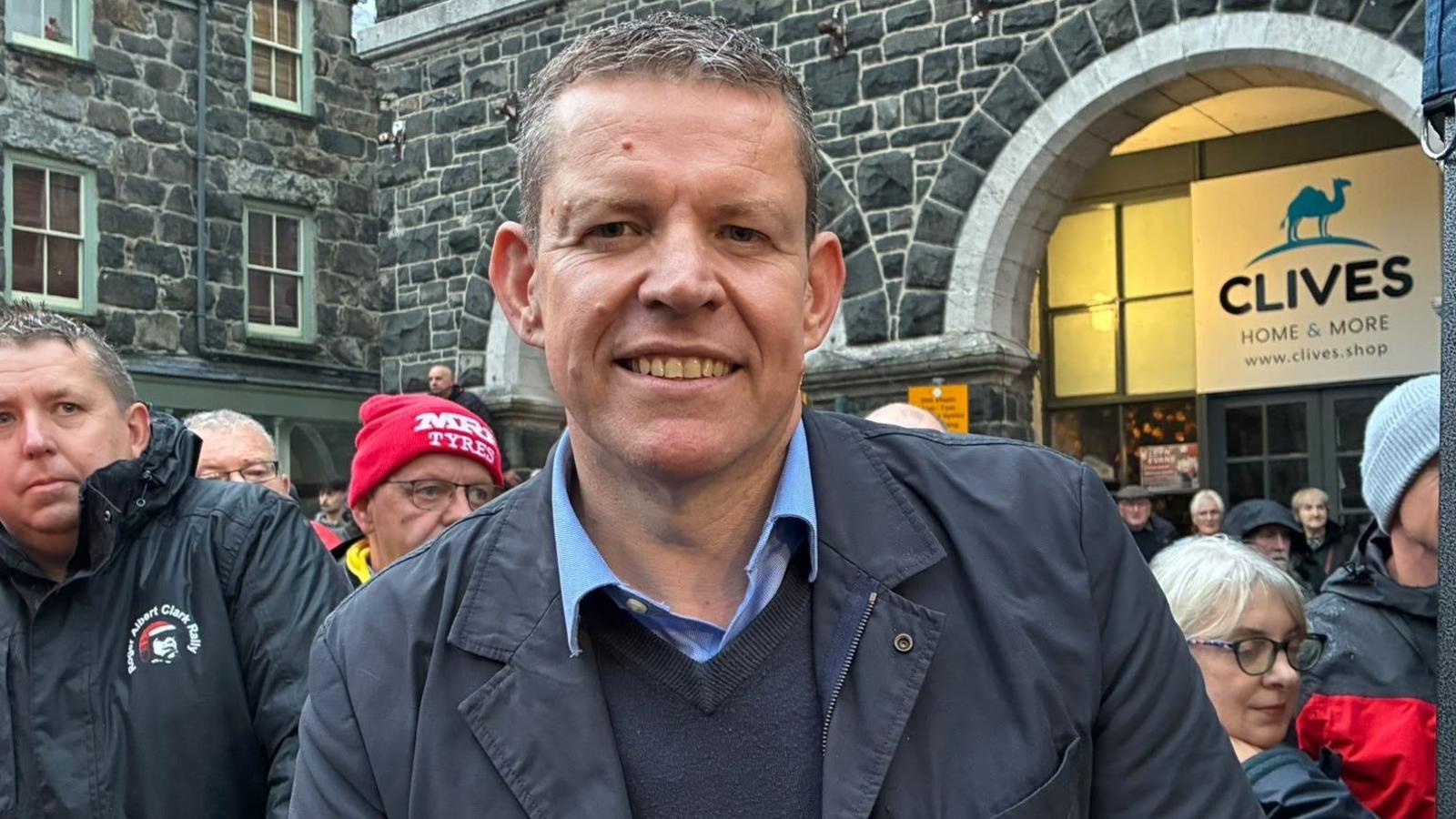 Rhun ap Iorweth on the sidelines among the crowd wearing navy jacket and navy v-neck jumper and light blue shirt. Grey stone buildings are visible behind him.