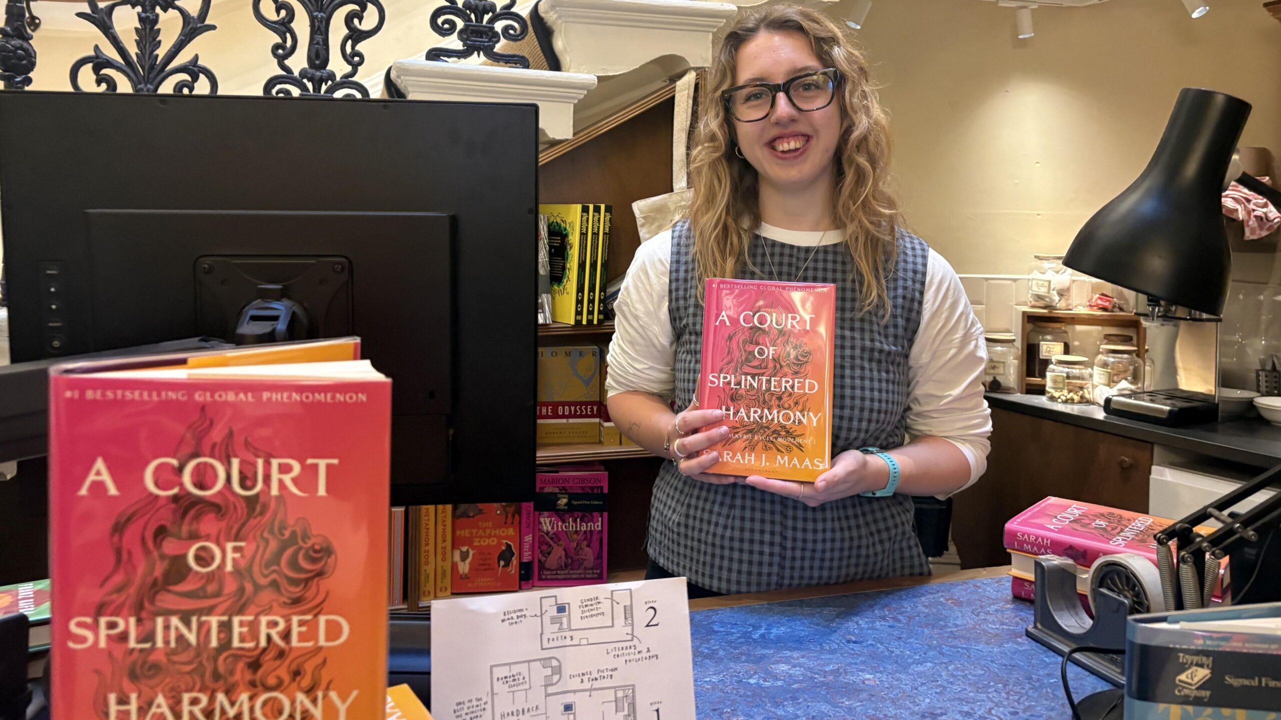 A woman with blonde hair and glasses is standing behind a counter, holding a book that reads: A Court of Splintered Harmony