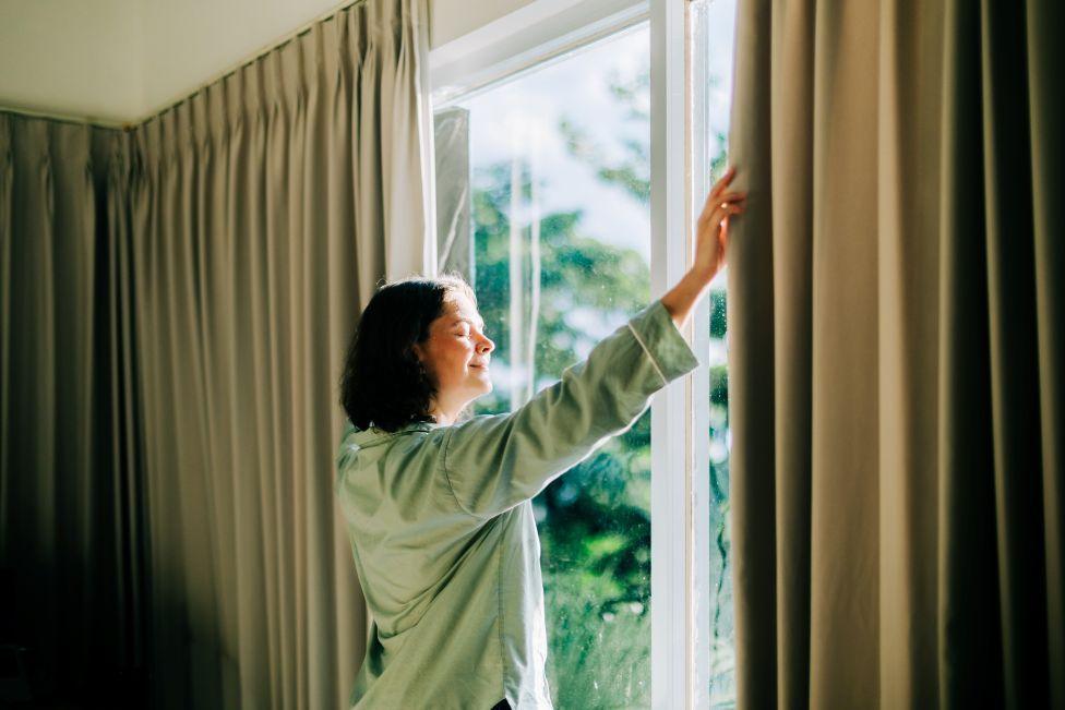 A woman in a green top opens curtains. Out of the window you can see lots of green trees
