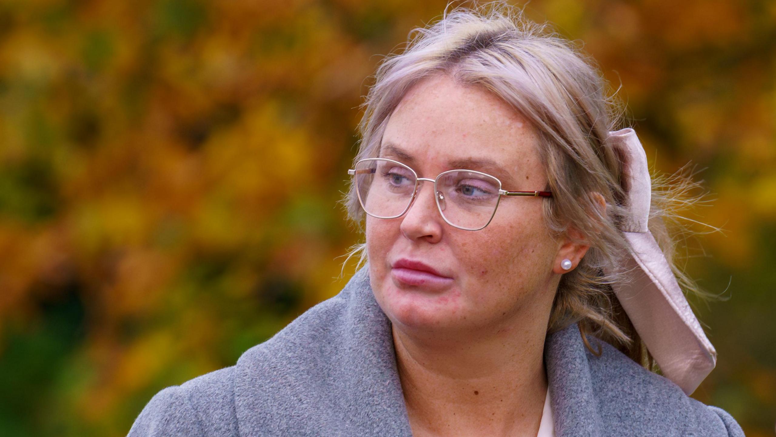 A blonde haired woman, with her hair tied back in a light pink bow. She wears gold rimmed glasses