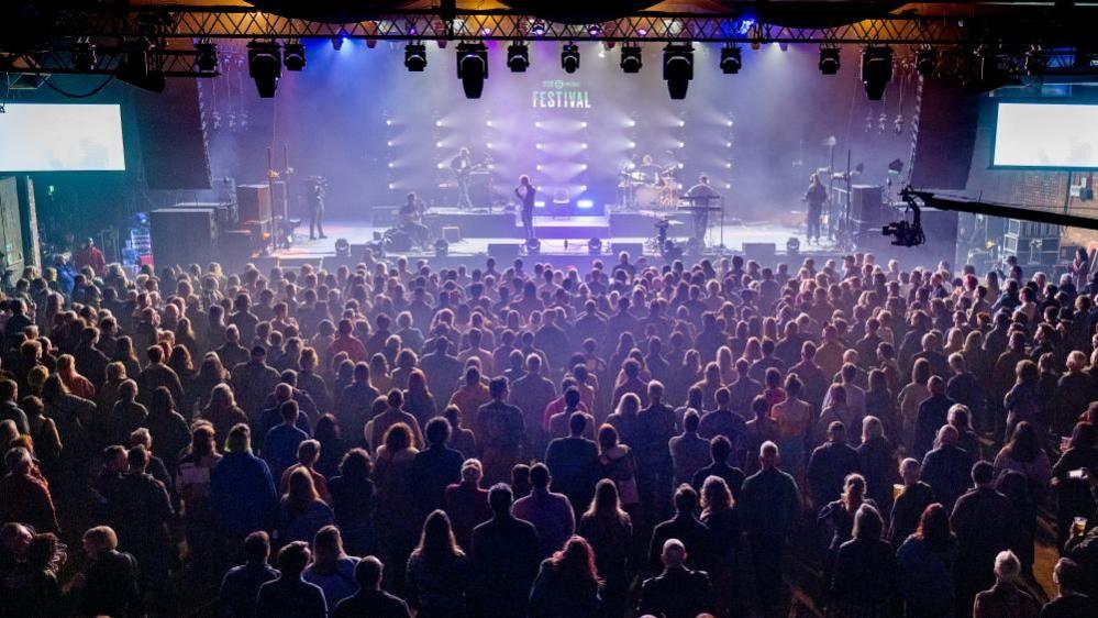 An image of a large crowd of people watching people perform on a stage in front of them take from above.