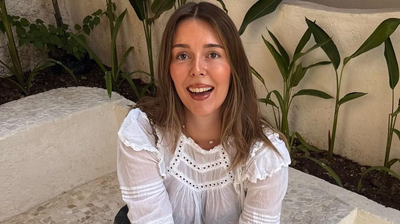 Young woman with long brunette hair smiling, wearing a white long-sleeved top and sitting on a wall, with green plants behind her.