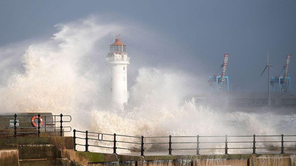 Large waves crashing against the sea wall at New Brighton promenade. A white lookout tower is in the middle of the image, partially obscured by the waves and spray. The sky is overcast.