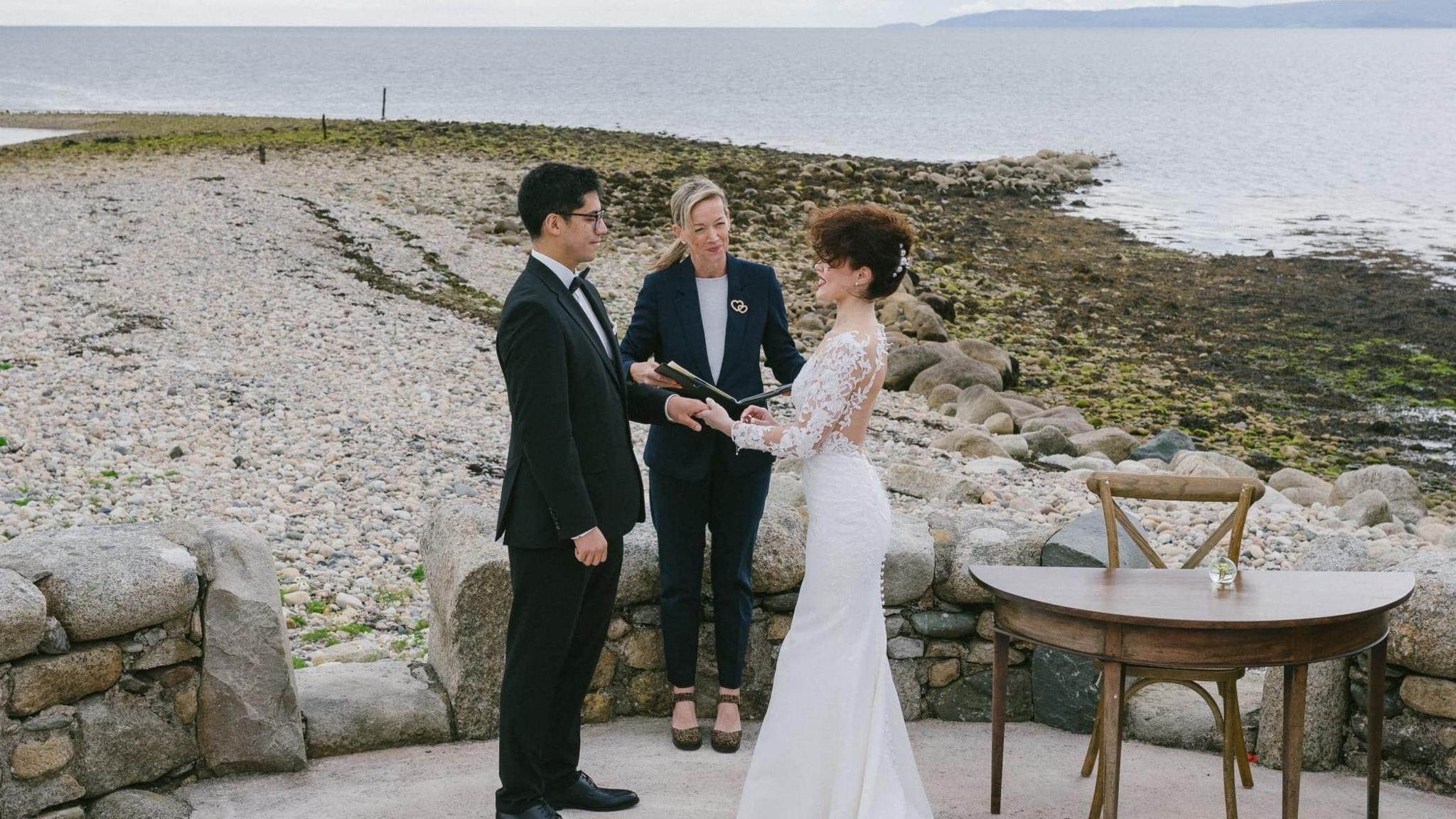 Chihon and Ana-Maria stand with an officiant as they say their vows. Behind them is a stony beach with sea and hills in the distance. Chihon has black hair and wears a black suit, across from him, Ana Maria has brown hair in an updo and wears a white lace wedding dress.