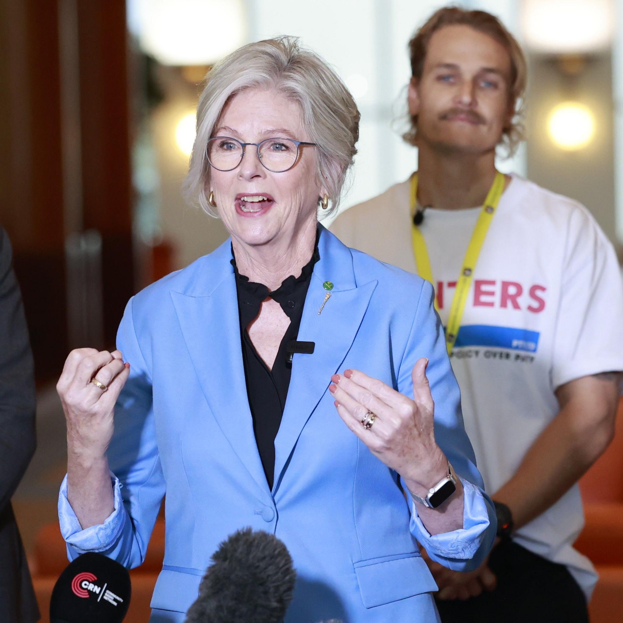 Helen Haines, wearing a light blue blazer and glasses, speaks to reporters indoors, gesturing with both hands. Several microphones are visible in the foreground, while a man wearing a white T-shirt and yellow lanyard stands blurred in the background.