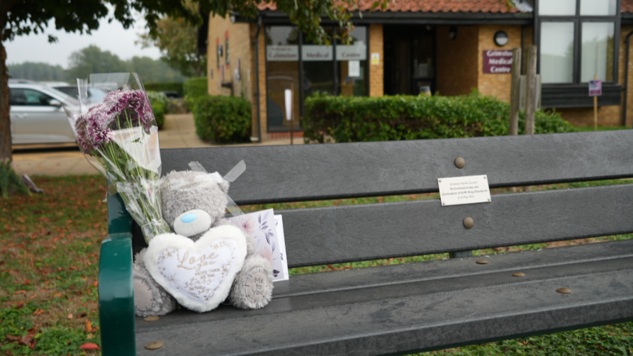 A grey teddy bear and flowers placed on a bench outside a doctor's surgery in Dersingham