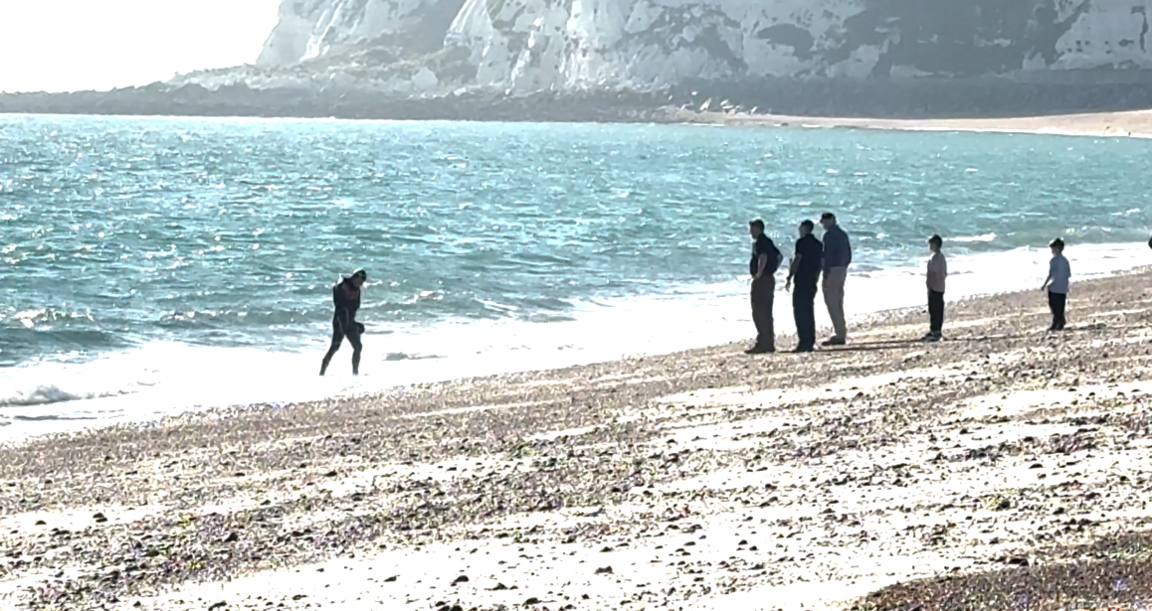A distance shot showing a man in a wetsuit wading out of the English Channel, the white cliffs of Dover in the background. Five people are on the sand to greet him.