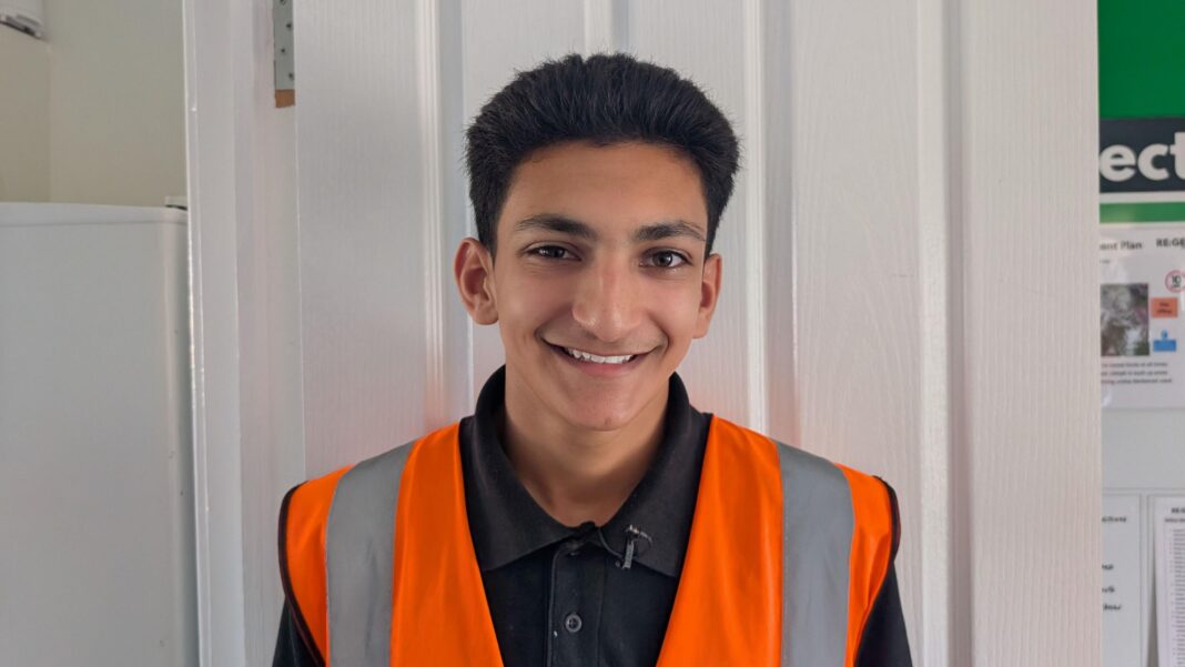 Param is pictured wearing a high-visibility orange safety vest over a black collared shirt, standing indoors in front of a white panelled door. Part of a noticeboard and a white appliance are visible in the background.