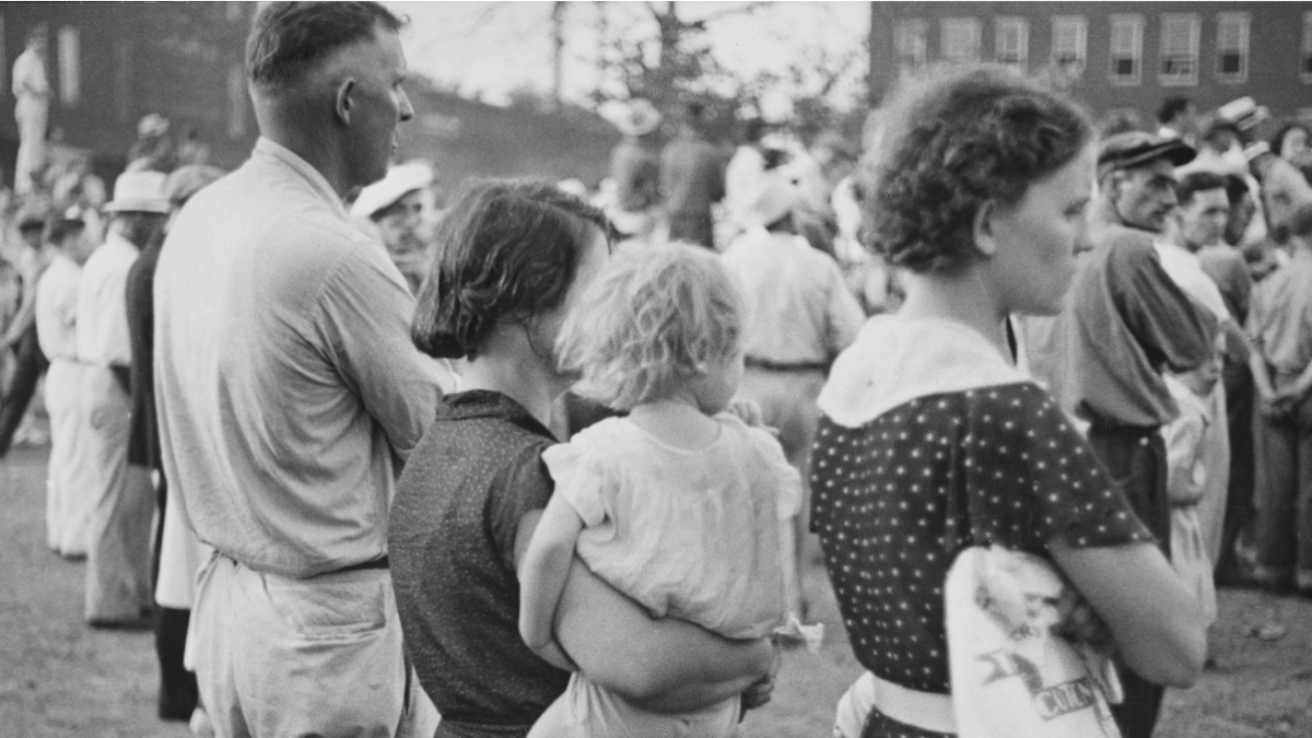 A woman in a dark collared dress holds a blonde child on her hip, standing between a woman in a polka dot dress and a man in light-coloured slacks and a shirt