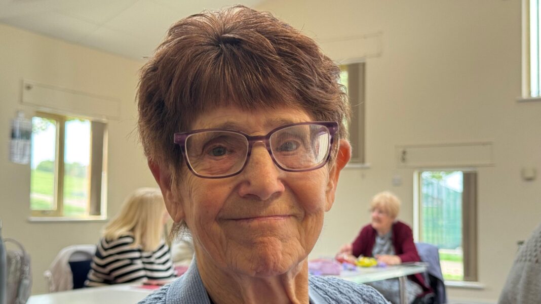 Tories committed to pension triple lock, deputy leader says An elderly woman with short brown hair and smiling is wearing glasses and a blue cardigan. Behind her are other pensioners, who are sitting around a table in a community centre.