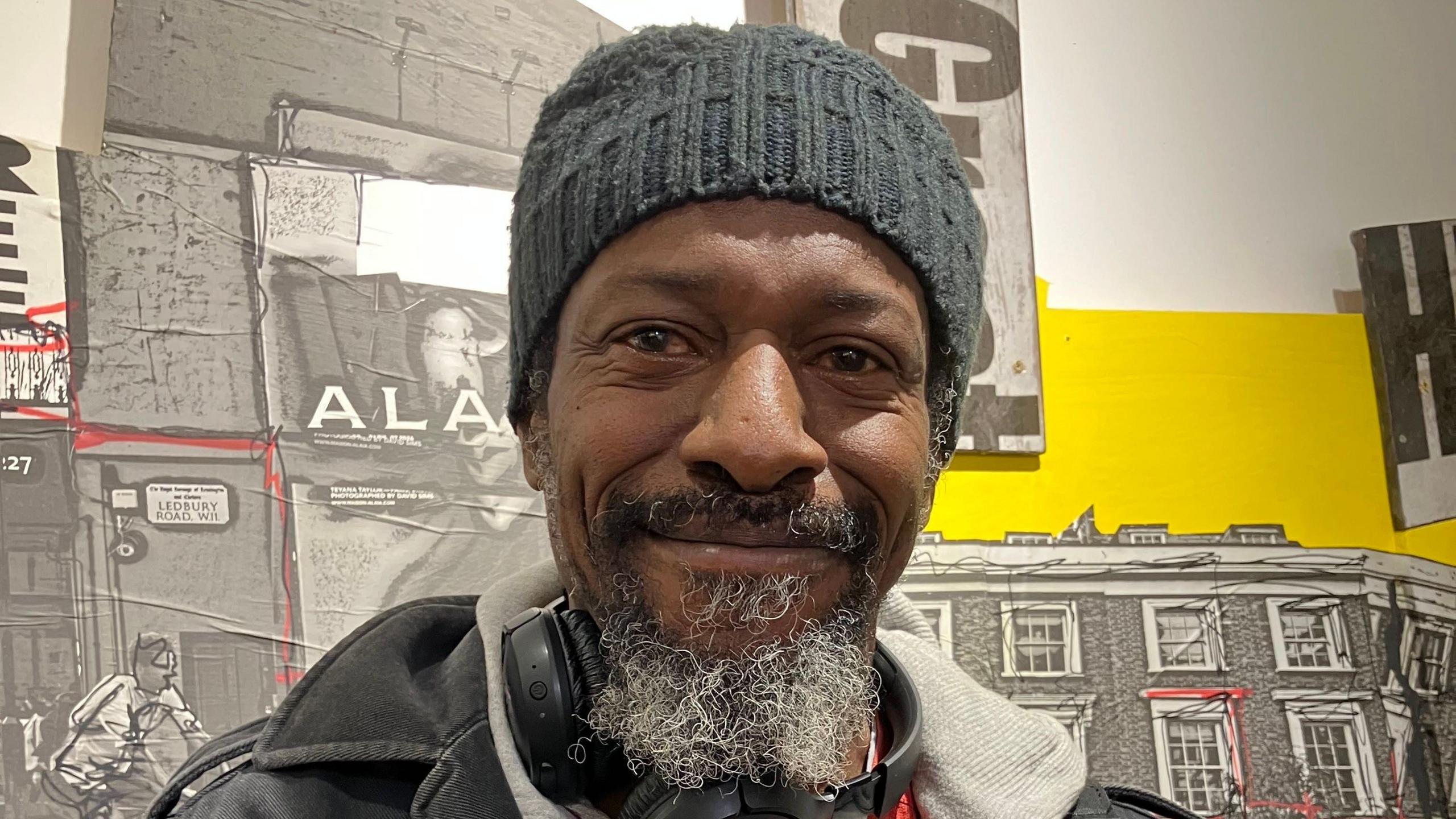 Karl Amata wears a knitted grey beanie, a dark jacket and grey hoodie. He carries a guitar and has headphone strung around his neck.