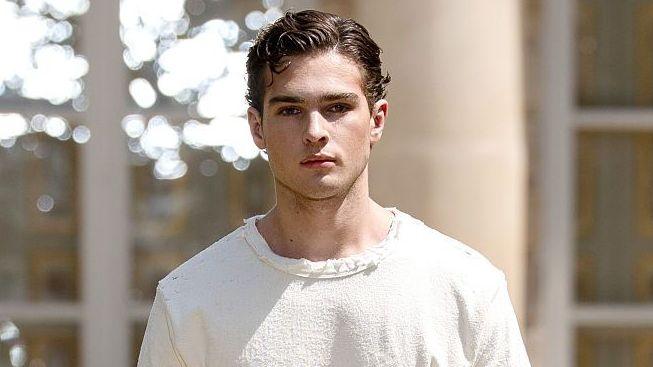 Close-up of Clavicular's face, looking serious, wearing a white sweater in front of white building