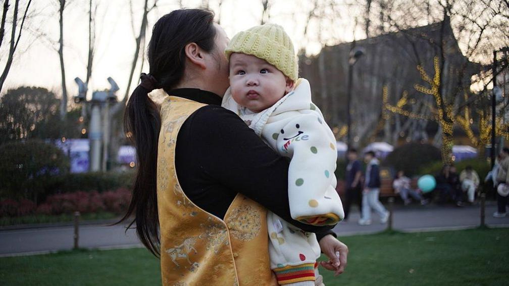 A woman in a golden vest holds a baby wrapped in a polka-dot blanket in the park at dusk