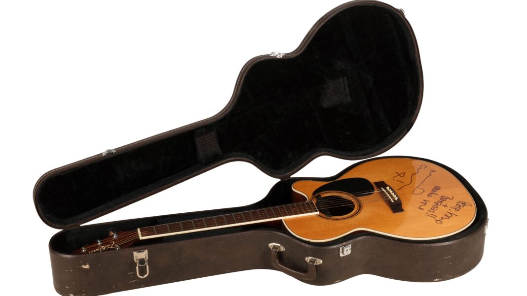 A signed guitar, in a brown guitar case. The wooden guitar has been signed by Noel Gallagher. It is on a white backing. The case is open.