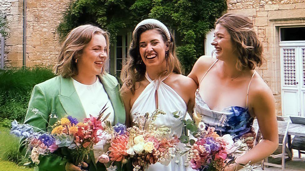 Lizzy, Charlotte and Libby picture at Charlotte's wedding. They are all holding bunches of flowers with their eyes closed as they smile. Charlotte is wearing a white dress, while Lizzy is wearing a light green suit jacket and Libby a multicoloured dress