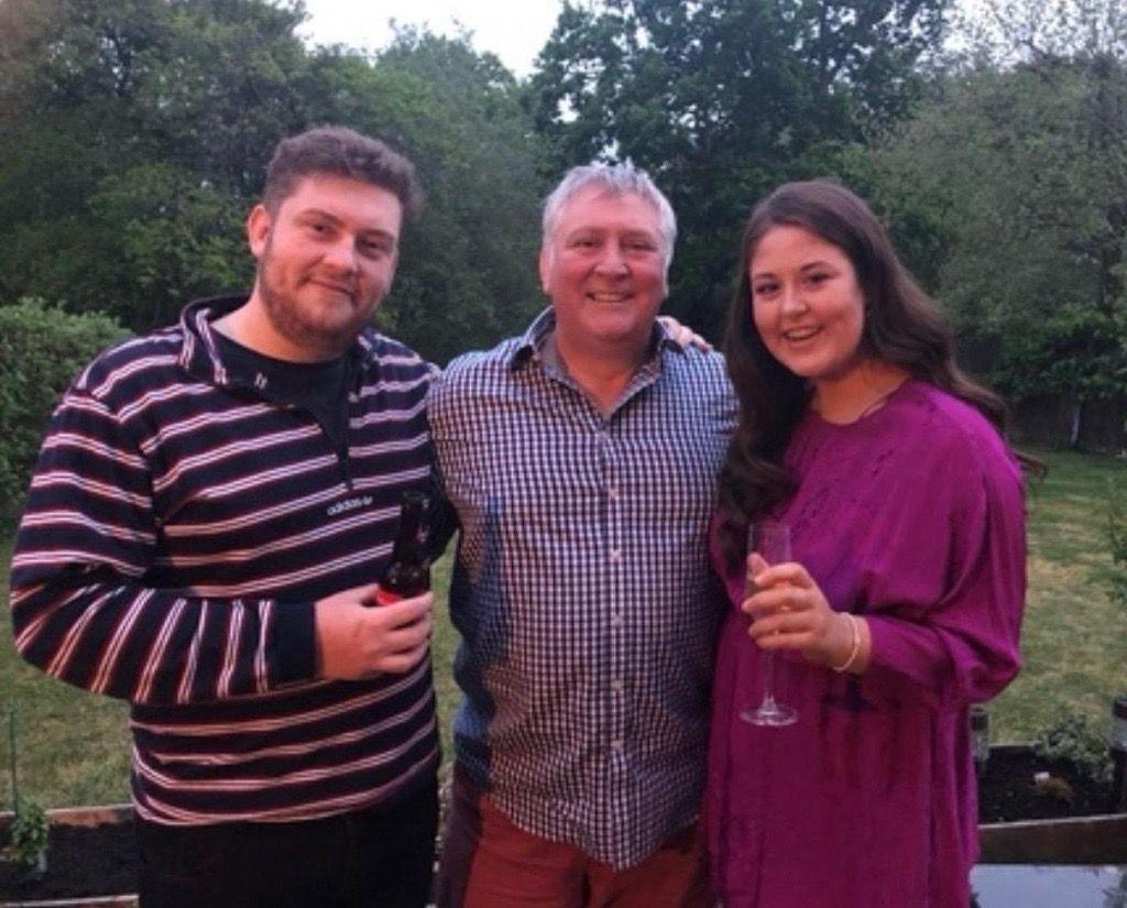 John standing in the middle with his arms around his son James on the left and daughter Amy on the right. They are in a garden and Amy is holding a champagne flute and James a beer.