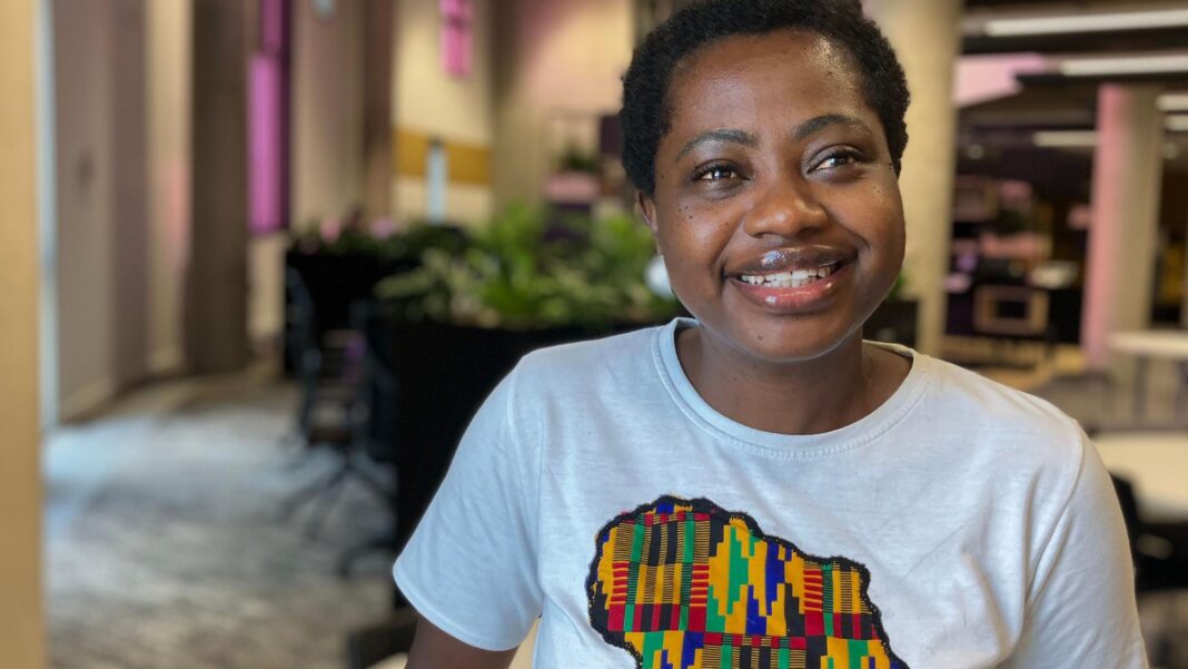 A woman stands smiling at the camera. She is wearing a T-shirt with a colourful design in the shape of a map of Africa.