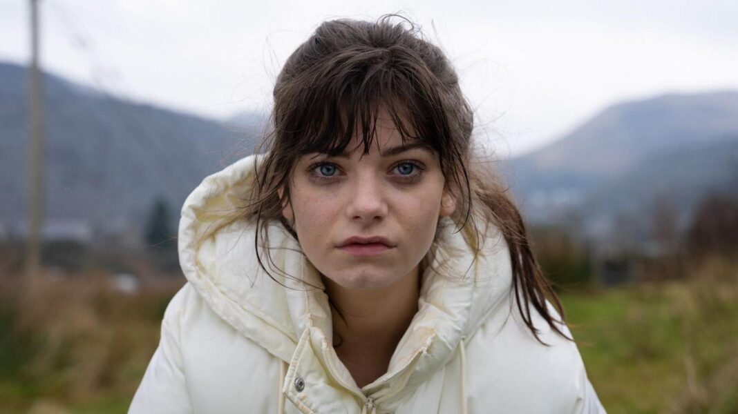 Effi, a young woman with brown hair in a ponytail and wearing a white puffer coat, stares at the camera with a sad expression.