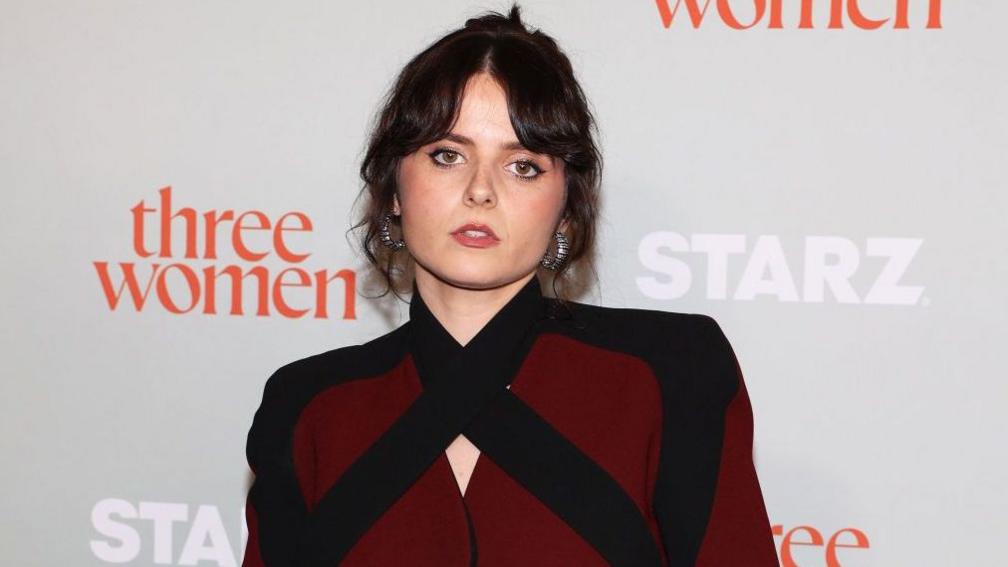 Gabrielle Creevy, on the red carpet. Her dark brown hair is tied back loosely and frames her face and she wears large silver hoop earrings and a burgundy and black top.