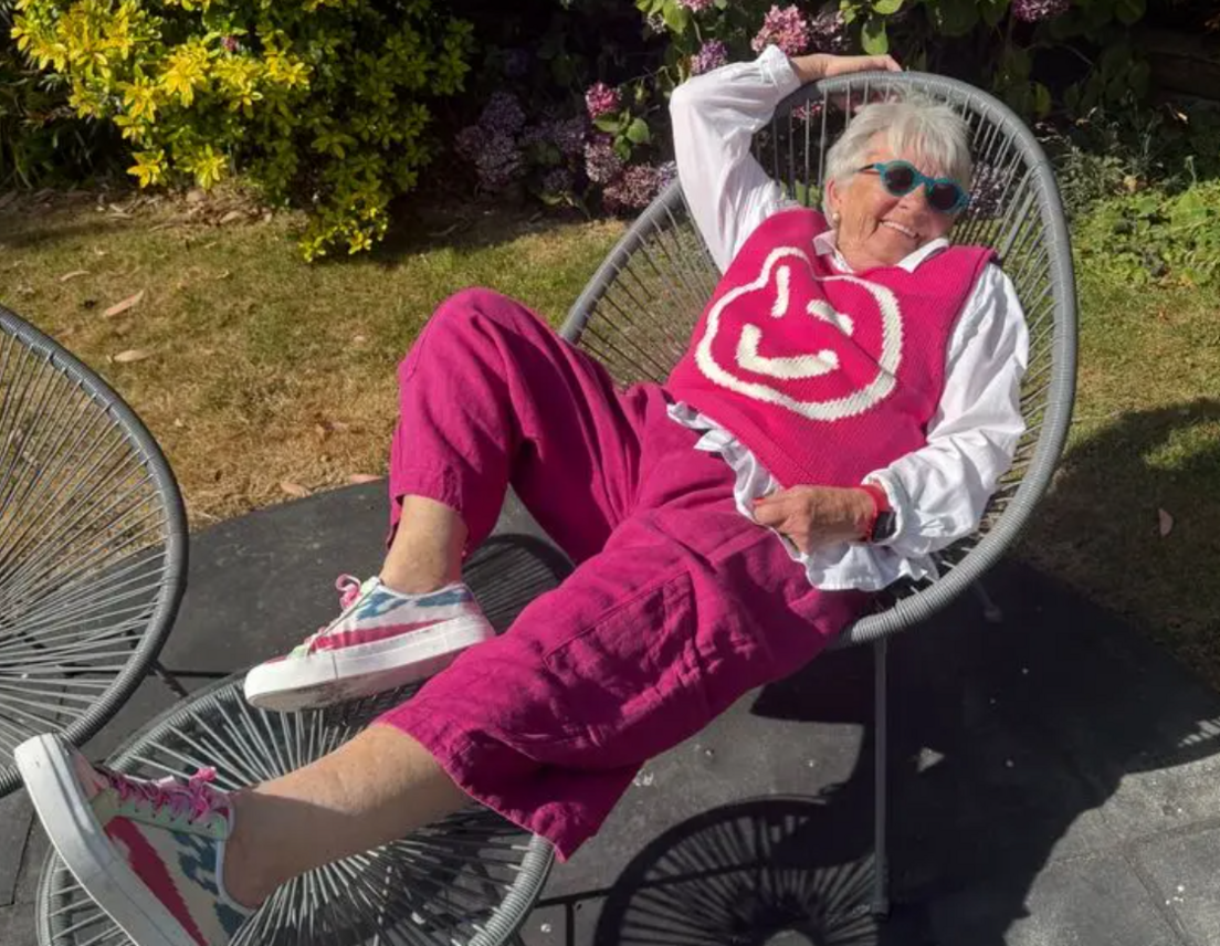 Viv Truran with grey hair, lying on a chair outside wearing a pink jumper and pink trousers