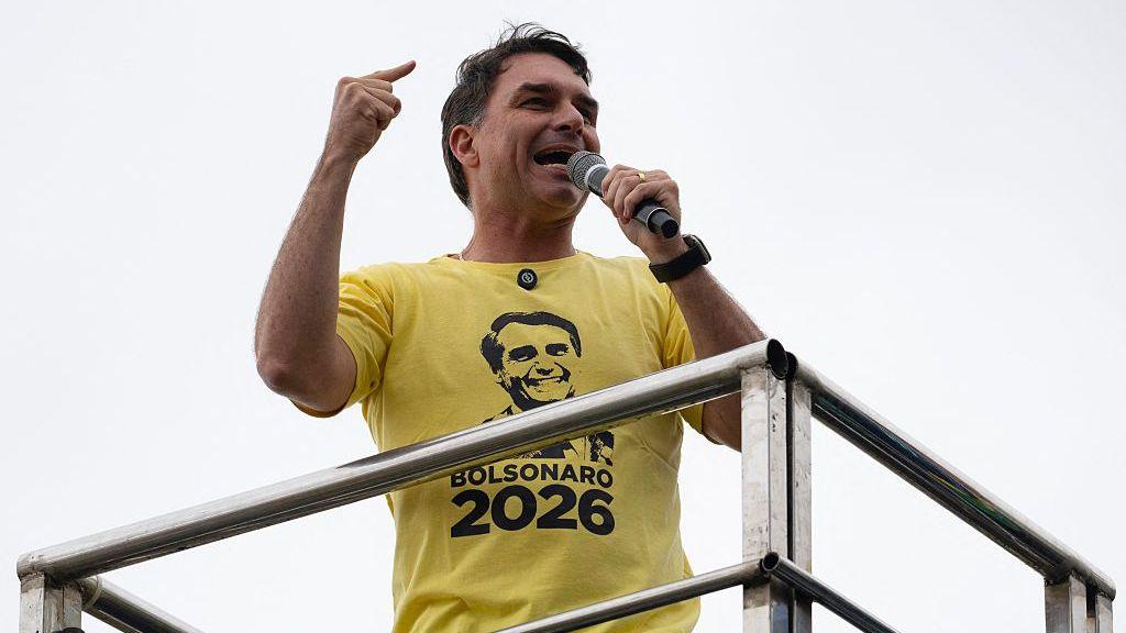 Flávio Bolsonaro gives a speech while wearing a yellow t-shirt with his father's face on it.