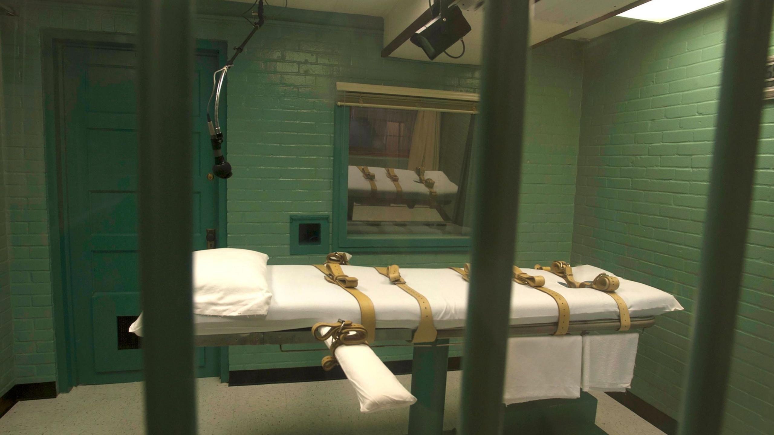 A gurney with sheets, a pillow and straps in a death chamber with a green door and green walls can be seen through the steel bars of the viewing area at the Texas state penitentiary in Huntsville
