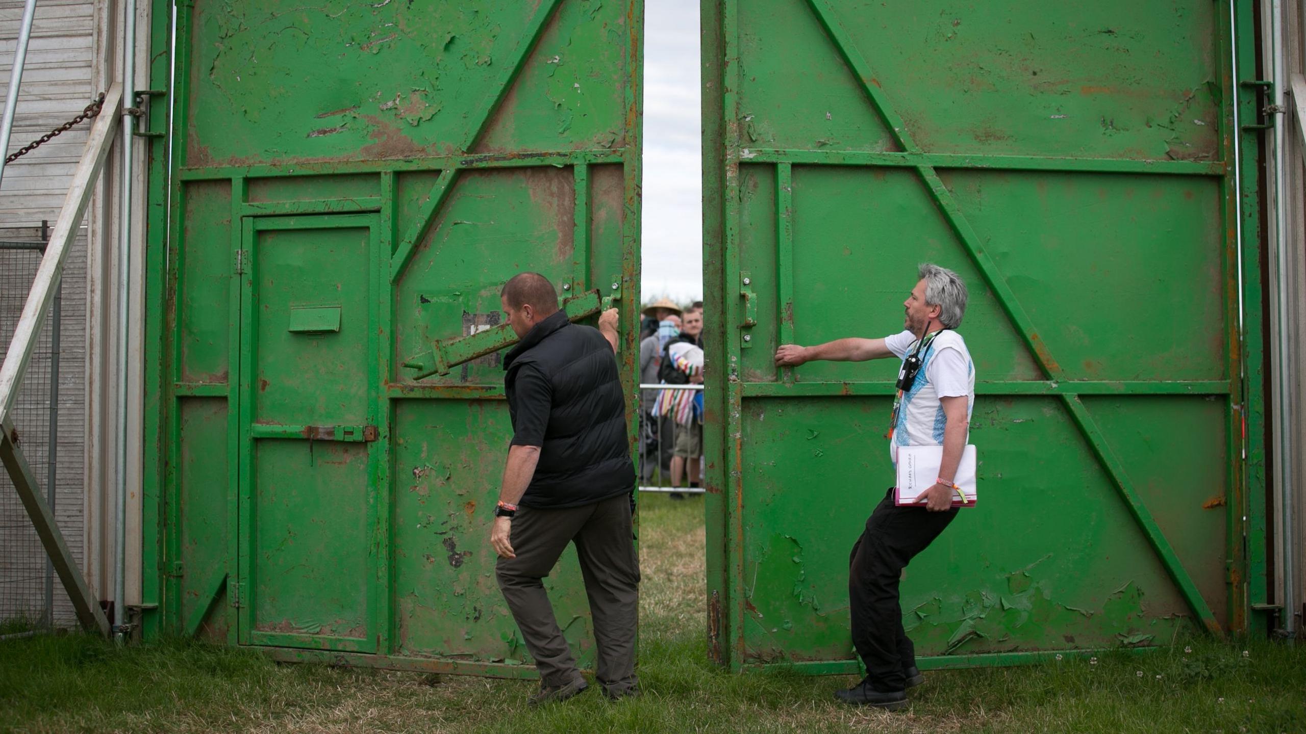Two men opening two green gates to Worthy Farm in Somerset for Glastonbury Festival. A large queue of people can be seen through the gap in the gates.