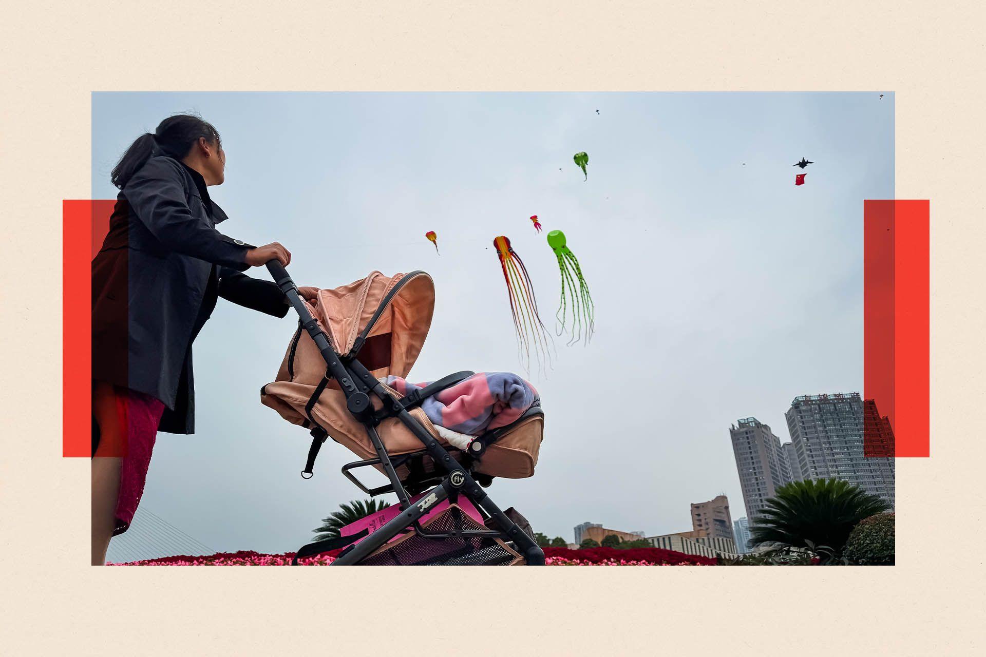 A woman pushes a pink pram near a city skyline. Two kites are flying in the sky