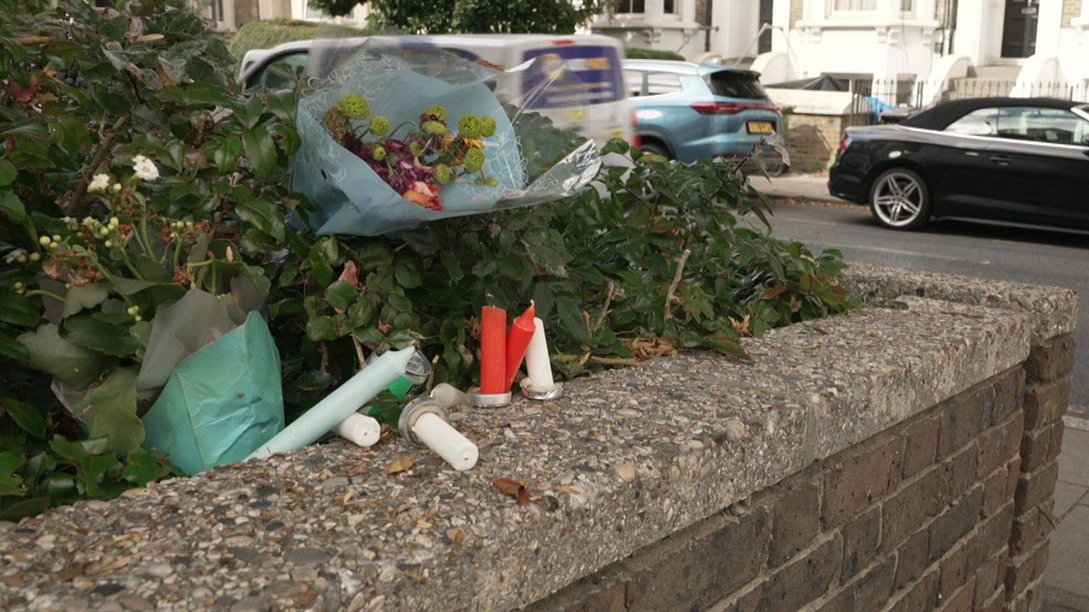 Two bunches of flowers, along with several candles, have been set up along the top of a wall on a roadside.