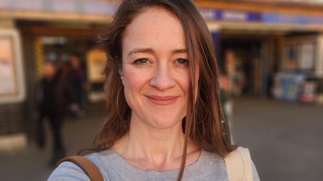 The singles choosing in-person dating events over apps Candice Gallagher with long brown hair and big hoop earrings smiles at the camera. A blurred London Underground station is seen behind her.
