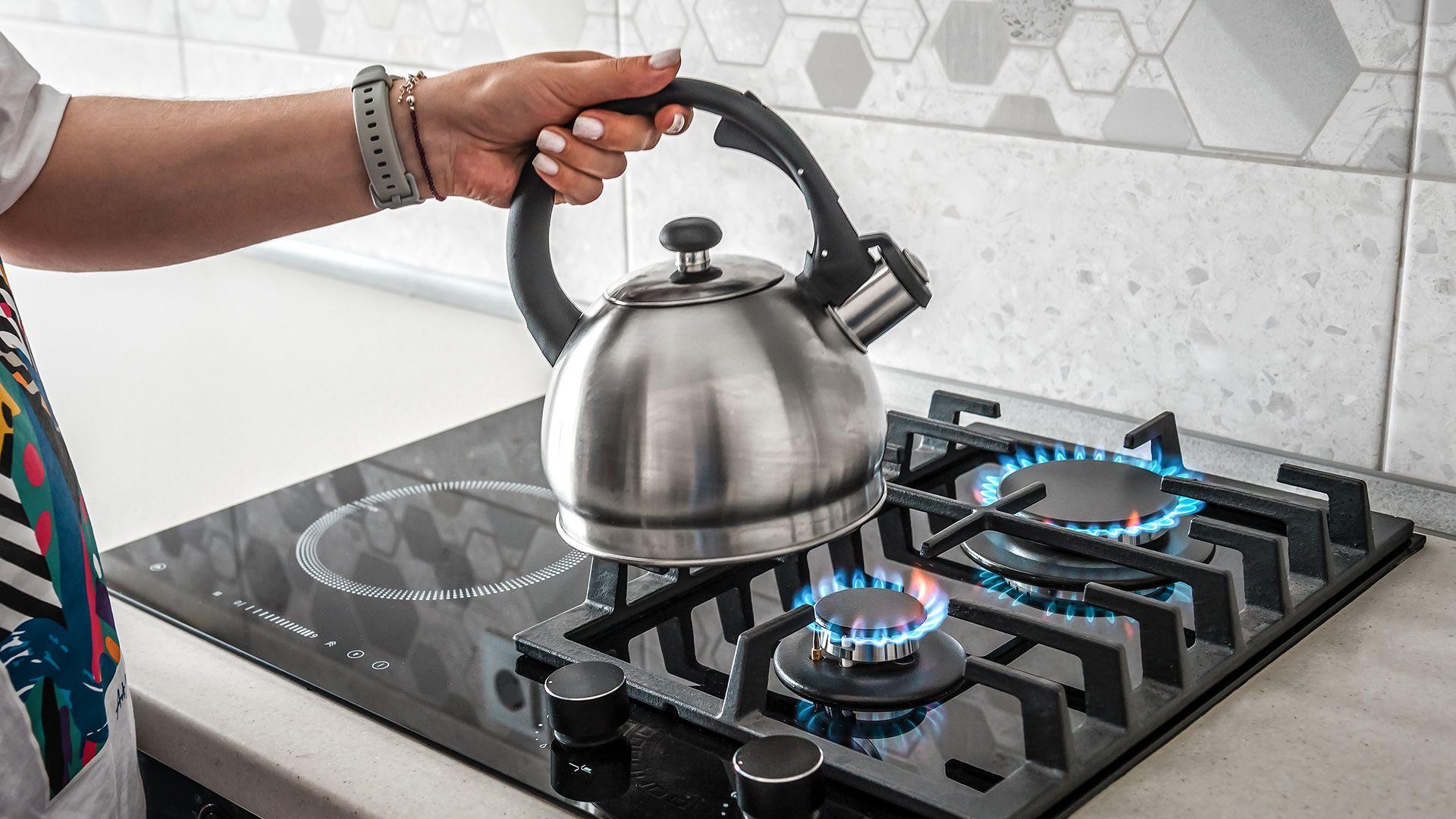 Stock photo shows a person's hand placing a kettle on a gas hob in a kitchen at home.