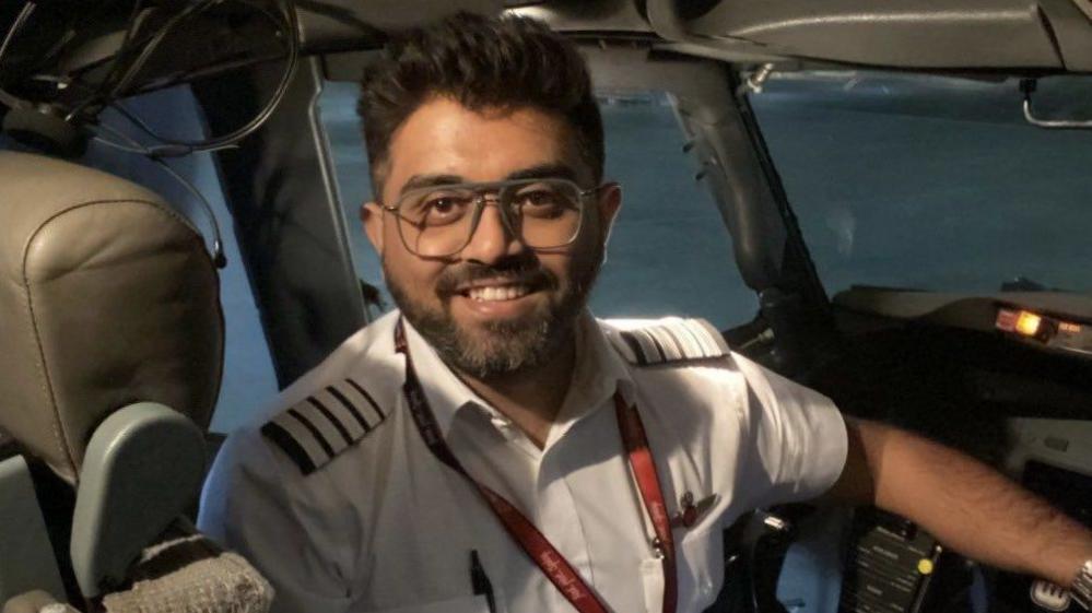 Indian pilot Smit Machchhar seen smiling into a camera as he sits in the cockpit of a plane. He is wearing glasses, a white shirt and has a pen tucked in his shirt pocket.
