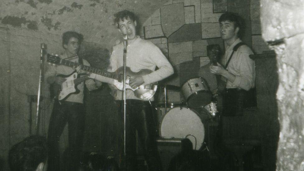 Black-and-white photograph of the Beatles performing on stage inside a small brick-vaulted cellar venue. Three musicians are visible with guitars and a drum kit, standing beneath the club's arched ceiling in front of a microphone, with audience members visible at the bottom of the image.