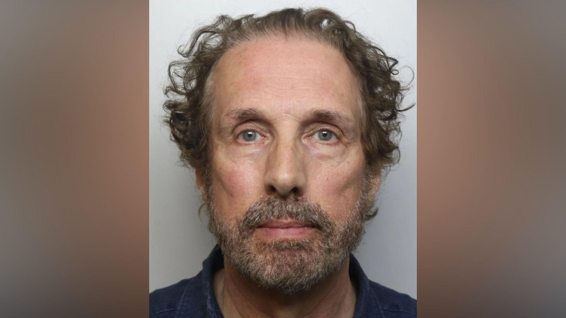 A police mugshot of Chris Brain. He has curly brown hair and a grey beard. He is wearing a navy blue shirt.