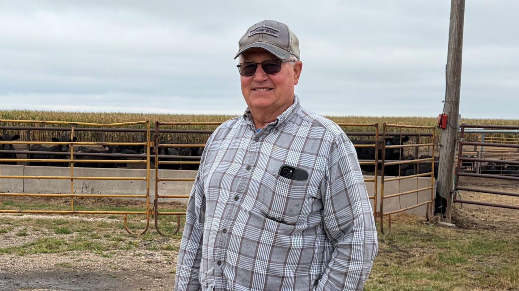 Don Cantrell wears a plaid shirt and a cap and stands in front of a farm fence