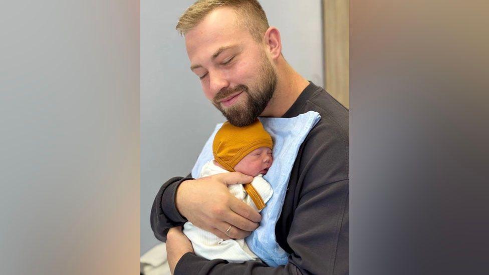 Nathan Stobbart is holding his newborn baby, who is wearing a white baby suit with a mustard-coloured hood and is fast asleep. Nathan has his eyes closed and is smiling. He has short blond hair and a darker brown neatly trimmed moustache and beard. There is a pale blue cloth or blanket between his chest and his baby. Nathan is wearing a dark grey, long-sleeved t-shirt.