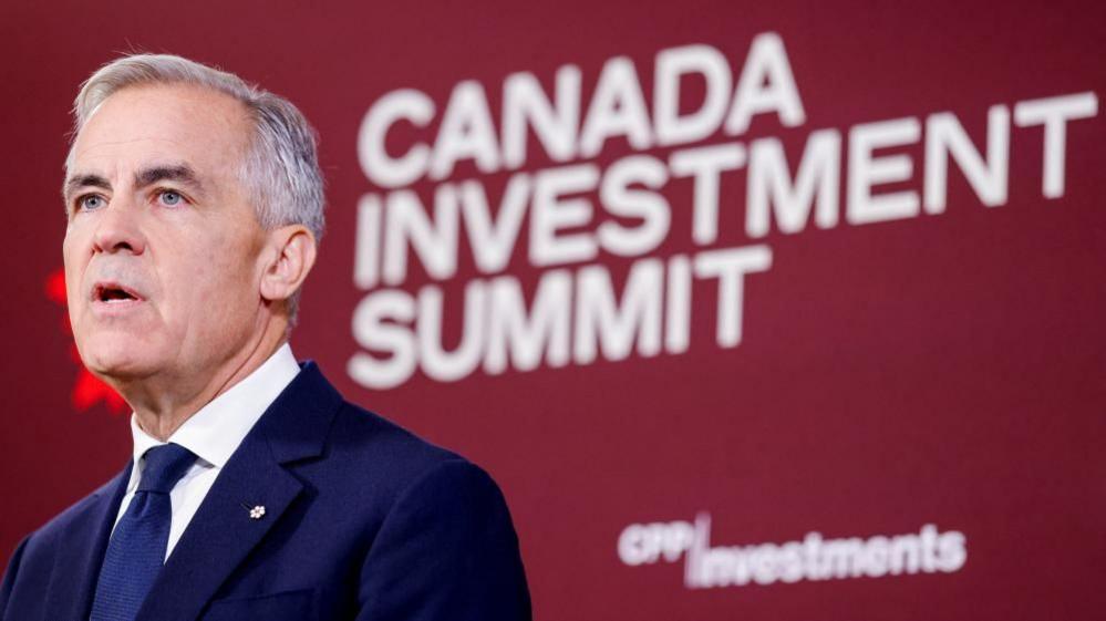 Mark Carney stands looking away in a dark suit and tie. Behind him is a maroon backdrop with the words "Canada Investment Summit"