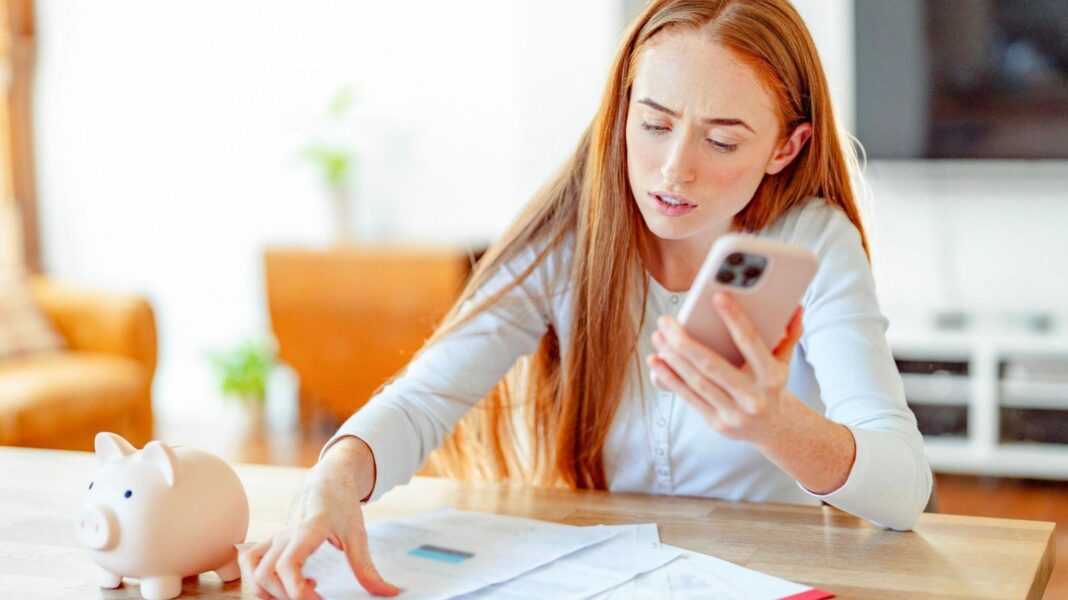 Woman with a phone in her hand and bills and a piggy bank on the table in front of her.