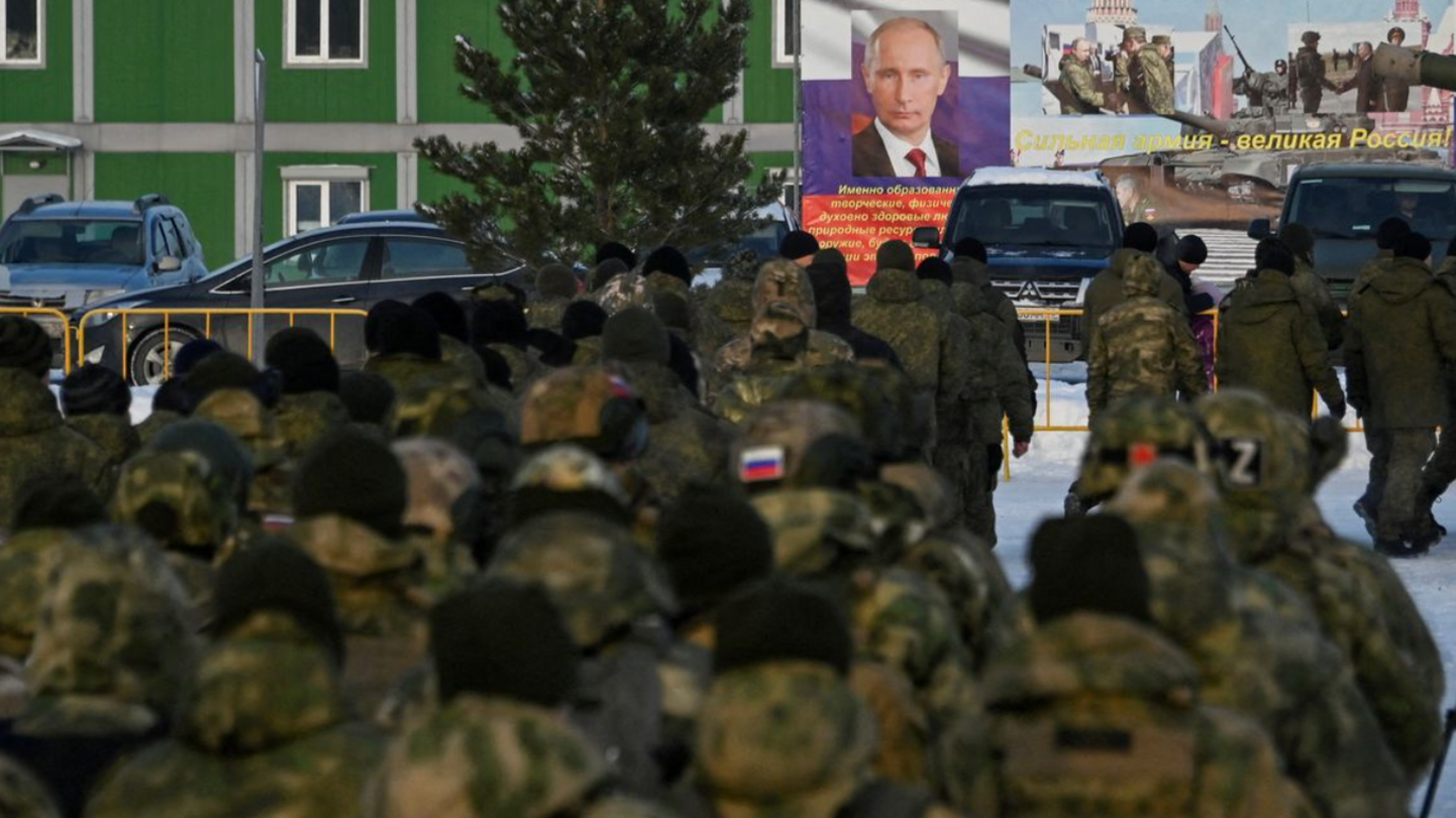 The backs of a crowd of soldiers wearing military camos with the Russian flag. They are facing a poster of Vladimir Putin