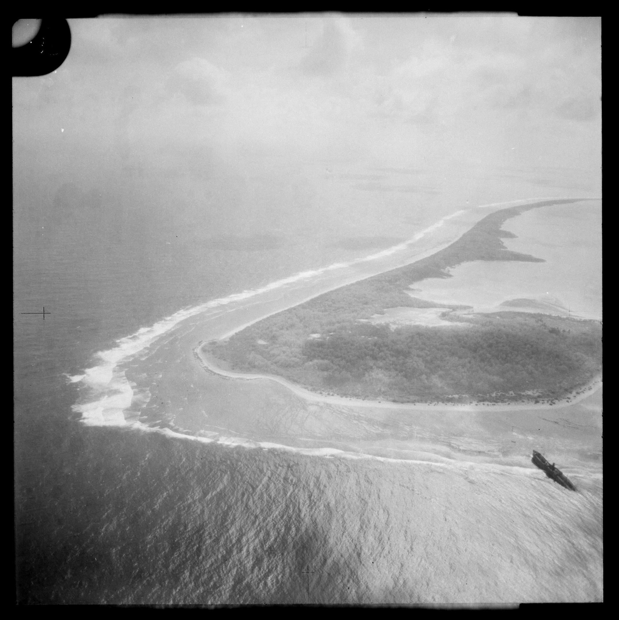 A black-and-white aerial image taken by a seaplane of Nikumaroro Island in the Pacific with the SS Norwich City wreck on its reef in the bottom right corner.