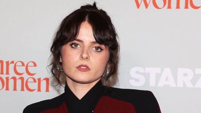Gabrielle Creevy, on the red carpet. Her dark brown hair is tied back loosely and frames her face. She wears large silver hoop earrings and a burgundy and black top.