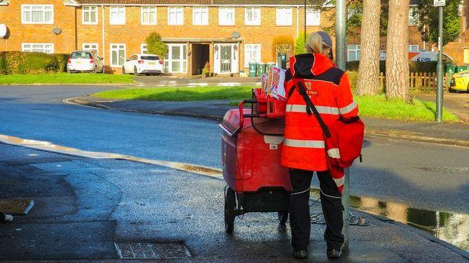 A postwoman seen from behind, standing wearing a Royal Mail coat and pushing a mail trolley on a residential road