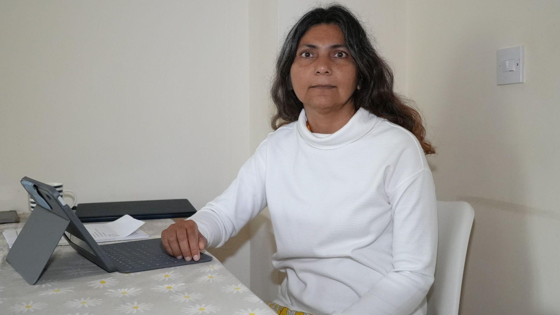 A woman with black hair and wearing a white turtle neck jumper. She is sitting on a white chair with an iPad with an attached keyboard. She is looking at the camera.