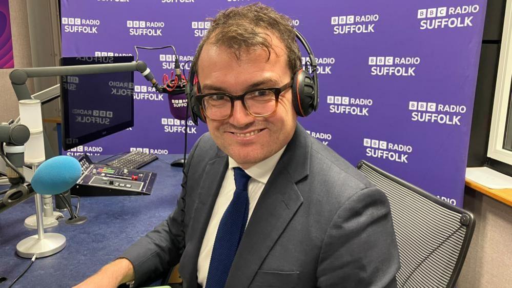 A head and shoulders shot of Patrick Spencer. He is sitting in front of a microphone in the BBC Suffolk studio and wearing headphones, a grey suit, white shirt, and navy blue tie. He is smiling into the camera.