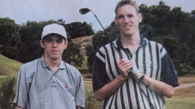 Two men stand side by side on a golf course, dressed in golf attire, with one holding a golf club across their shoulders. The photo appears to be an older snapshot, showing a grassy fairway, a path, and tree-lined scenery in the background under an overcast sky.