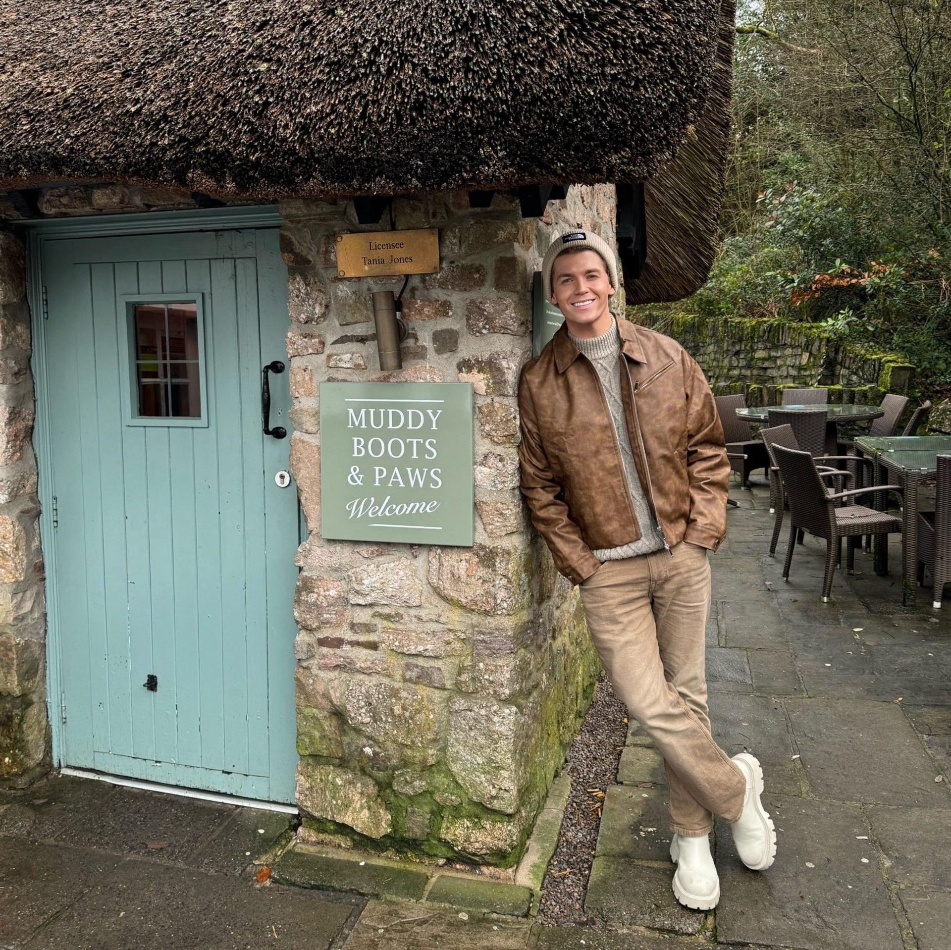 A young man wearing a beanie hat, grey jumper, brown jacket, brown chinos and white boots. He is leaning on the stone wall of a thatched building with a blue door and a sign that reads "muddy boots and paws welcome". He is smiling and looking at the camera.